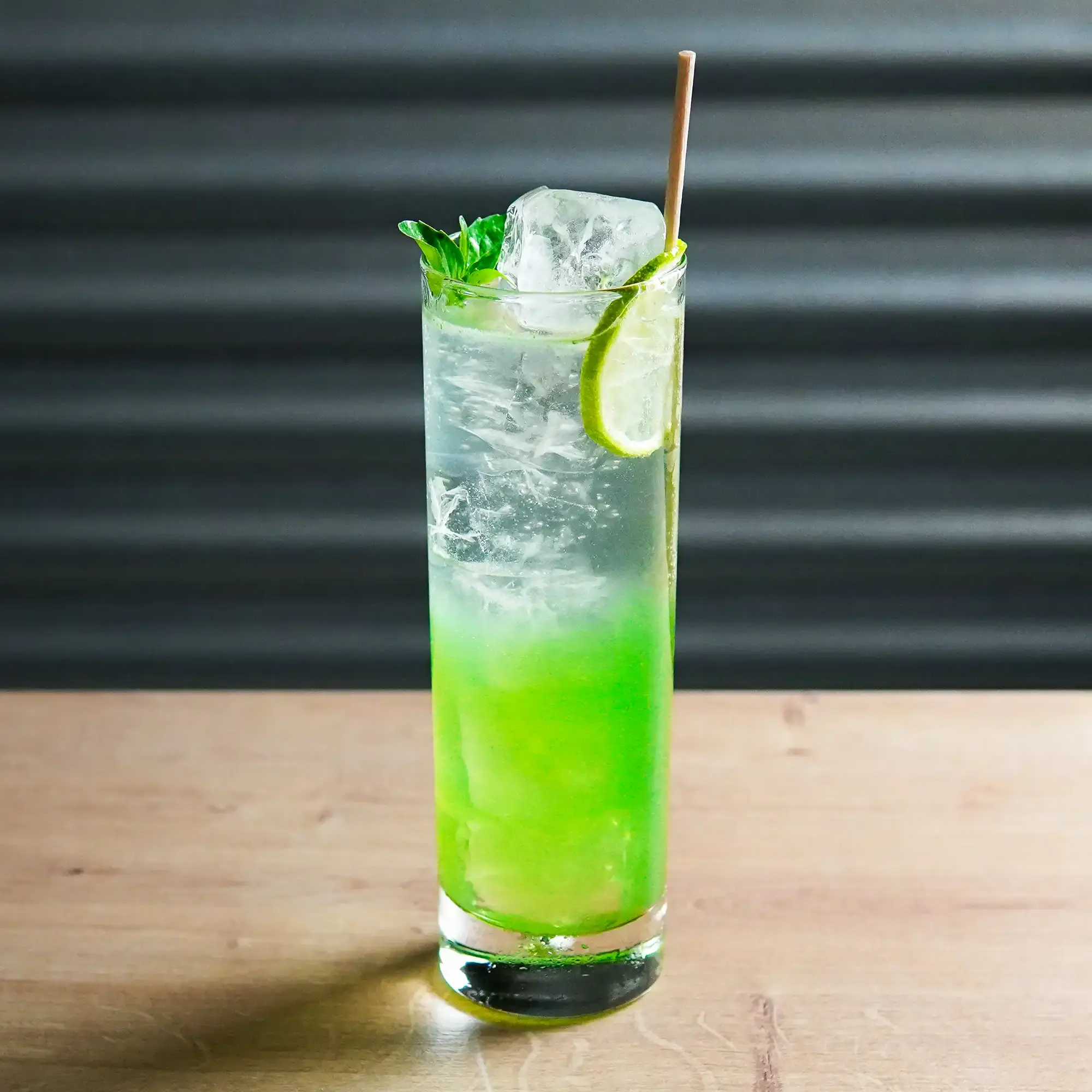 The image shows a tall, narrow cocktail glass on a smooth surface, illuminated by subtle background lighting. The drink has a striking color gradient, with a bright green layer at the bottom that transitions to a clear, transparent layer at the top. Large ice cubes fill the upper part of the glass, keeping the drink cool. A thin lime slice is attached to the rim of the glass, alongside a fresh green basil sprig. A slender straw, possibly made of bamboo or paper, extends from the glass. The background is dark gray with a subtle ripple texture, which emphasizes the bright drink in the foreground.