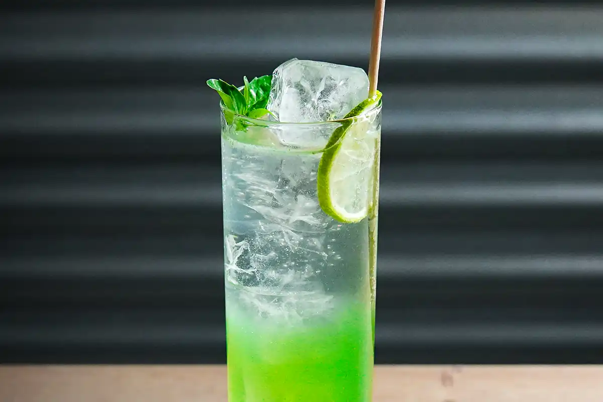 A tall glass of refreshing green cocktail garnished with a lime slice, basil leaves, and a large ice cube, with a straw on the side.