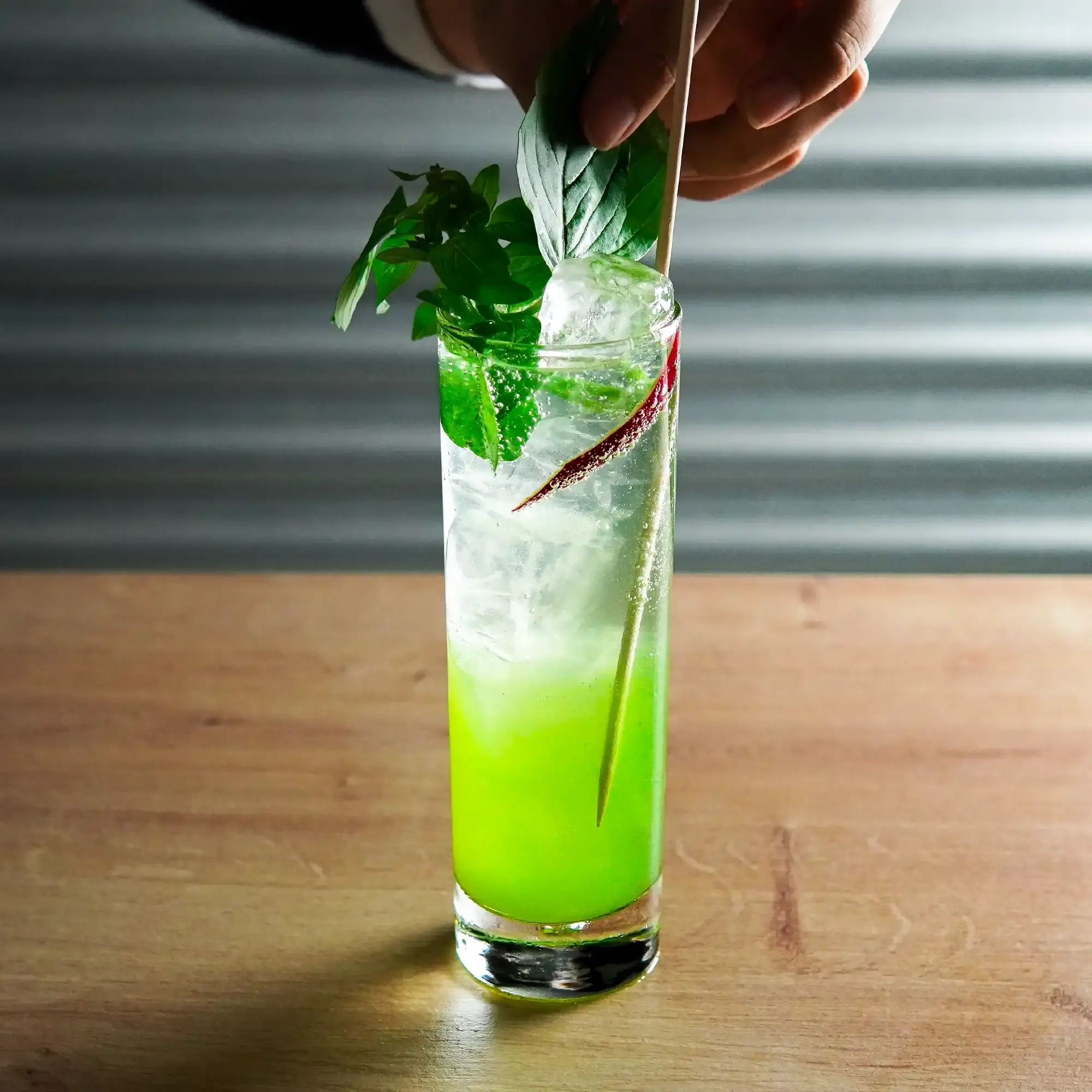 The image shows a tall, narrow cocktail glass placed on a light wooden surface. The drink in the glass has a vivid color gradient, with a bright green layer at the bottom, transitioning to a clear layer at the top. The glass is filled with large, clear ice cubes that create a refreshing appearance. A hand is seen reaching from above, carefully placing or adjusting a sprig of fresh green basil leaves as a garnish in the glass. Beside the basil, there is a thin, red garnish (possibly a chili pepper) adding a splash of color. A slender straw extends from the glass.