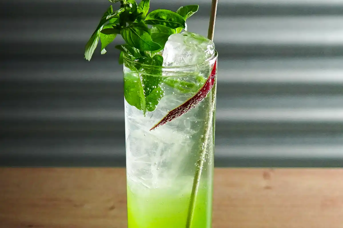 A tall glass filled with a vibrant green drink, garnished with fresh basil leaves, a large ice cube, a red chili slice, and a straw.