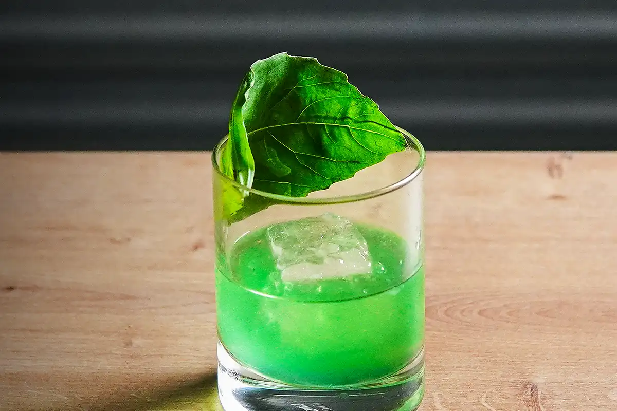A short glass with a bright green cocktail, topped with a large basil leaf and a single ice cube.