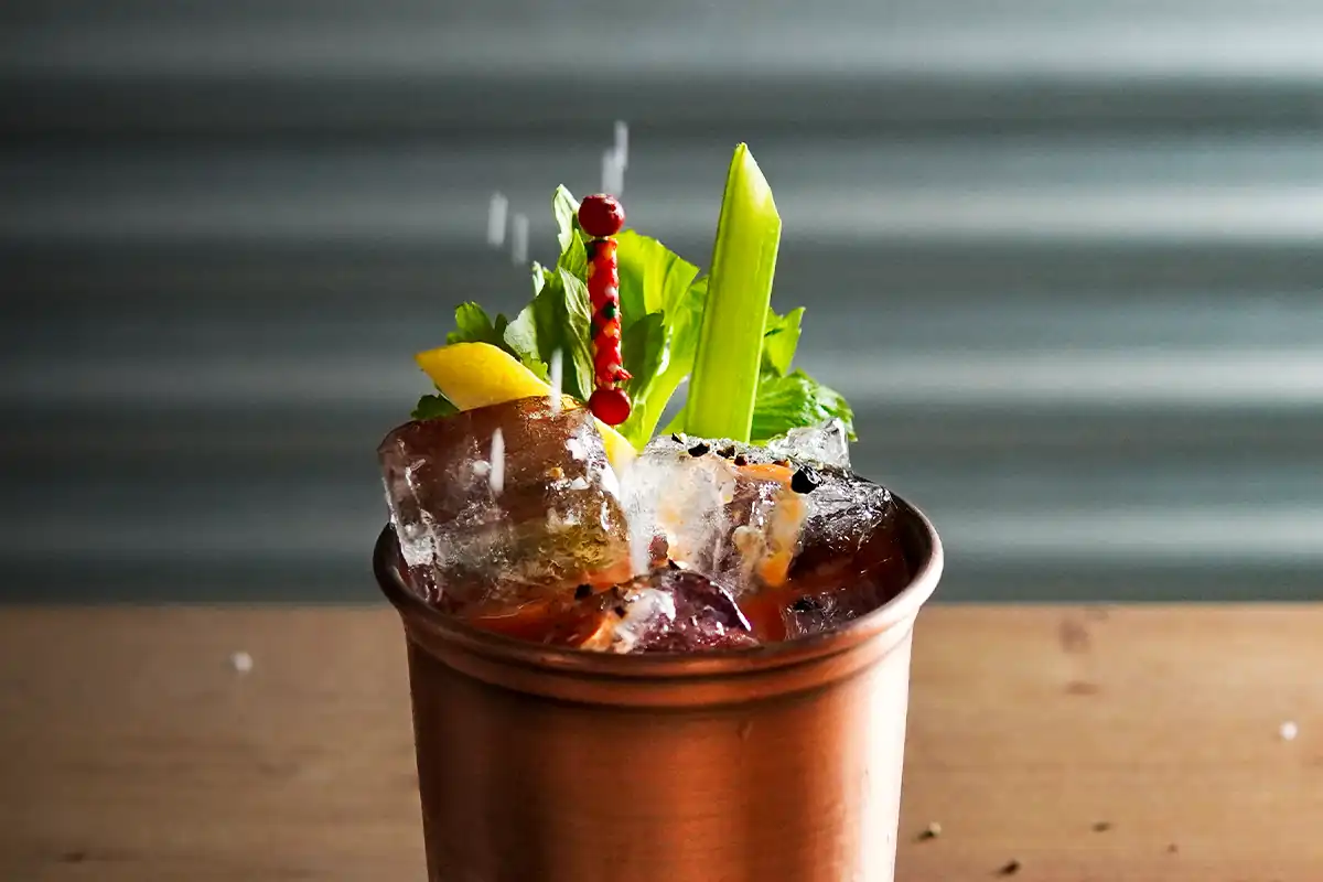 A copper cup filled with a vibrant cocktail, garnished with large ice cubes, a celery stalk, a lemon wedge, peppercorns, and a decorative red skewer.