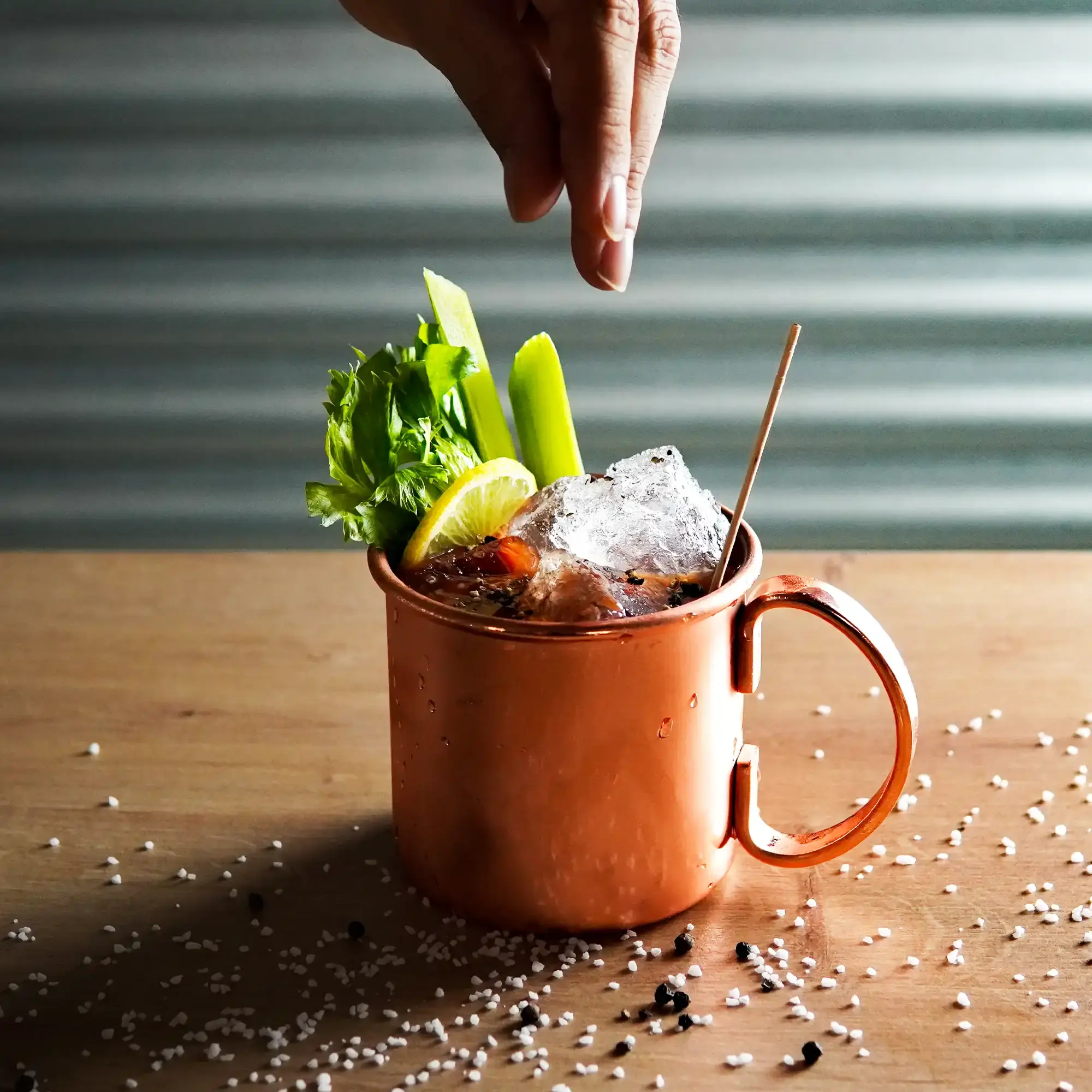 The image shows a copper-colored cup sitting on a light wooden surface. The cup is filled with ice cubes that extend above the rim. Various decorative garnishes are visible: a long red chili, a yellow lemon or lime zest strip, fresh green leaves (likely celery or herbs), and small black and white grains that resemble spices, scattered around the table. Above the cup, a hand is seen sprinkling a pinch of salt or spices over the drink, adding a dynamic touch. The background is dark gray and subtly textured, making the shiny copper cup and drink details stand out.