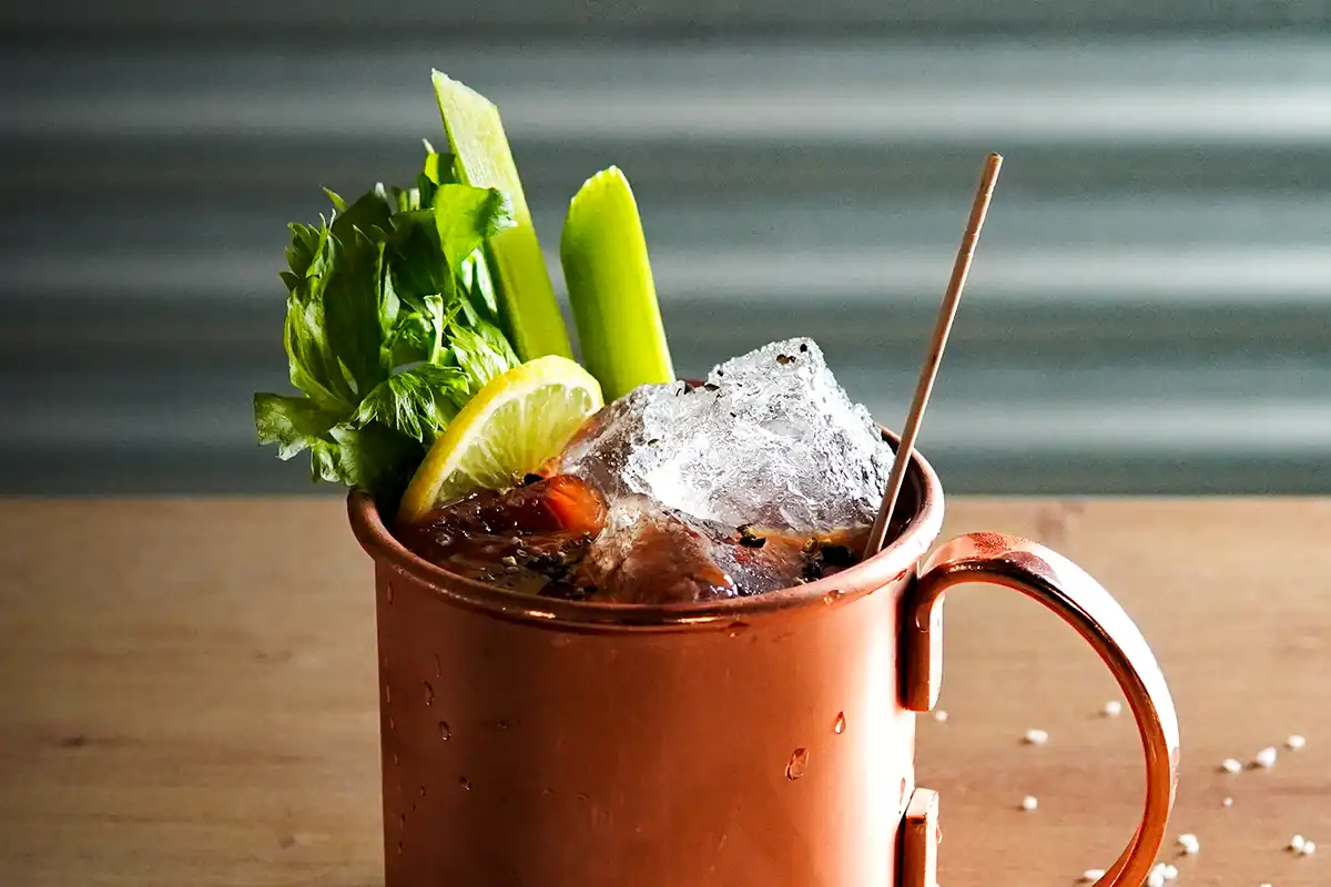 A copper mug filled with a dark cocktail, topped with large ice chunks, celery stalks, a lemon wedge, leafy greens, and a straw.