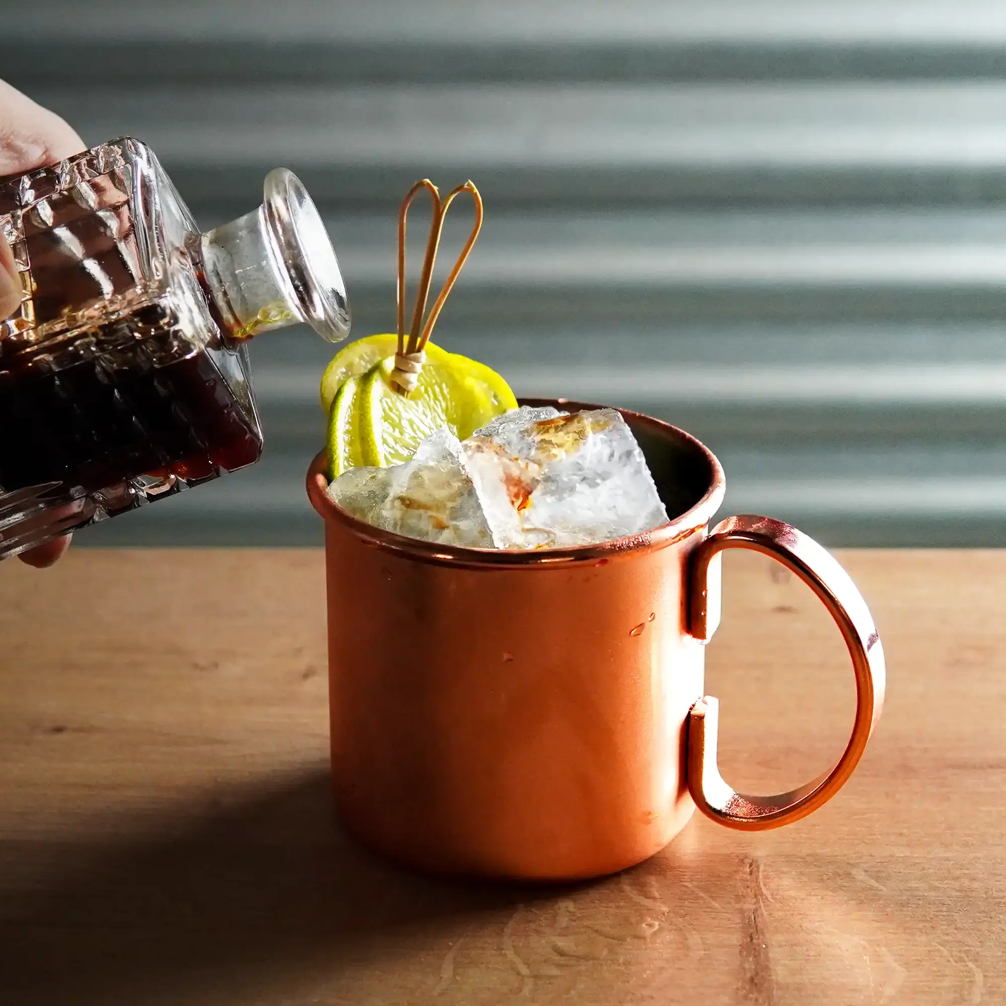 The image shows a copper-colored cup with a handle, sitting on a light wooden surface. The cup is filled with large, clear ice cubes that nearly reach the rim. Two lemon slices are attached to the side of the cup, held in place by a decorative pick. On the left side of the image, a hand is seen tilting a square, textured glass bottle and pouring a dark liquid into the cup. The bottle has a transparent design that emphasizes the dark color of the liquid. The background is gray and subtly textured, allowing the shiny copper cup and the details of the pouring liquid to stand out in the foreground.