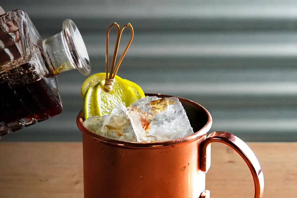 A copper mug with large ice cubes and lemon slices, as a dark liquid is being poured in. Garnished with a decorative skewer.