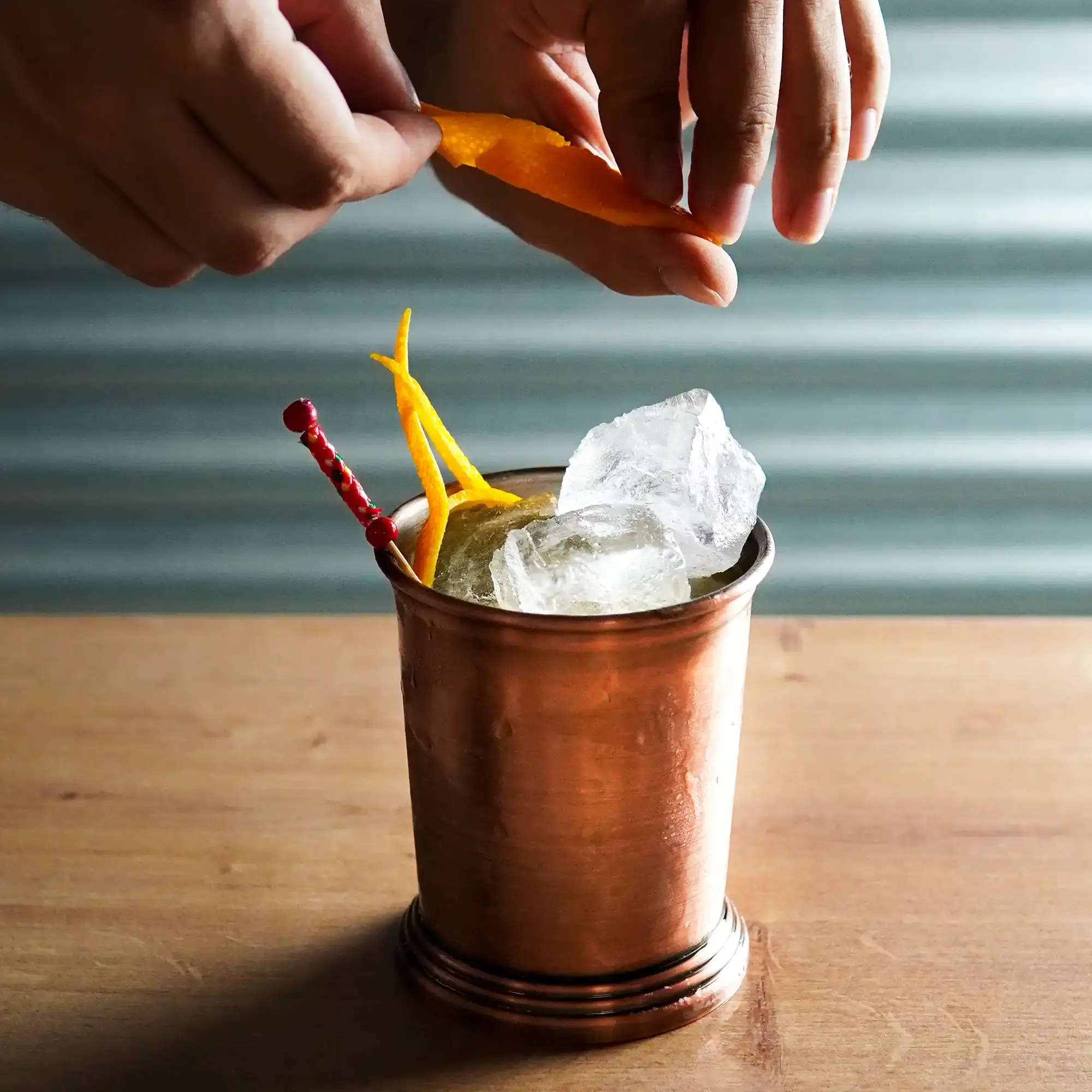 The image shows a copper-colored cup placed on a light wooden surface. The cup is filled with large, clear ice cubes, slightly protruding above the rim. Two long, thin strips of orange citrus peel are arranged decoratively in the cup. A red cocktail pick with a pearl-like appearance is also inserted into the drink, adding a splash of color. Above the cup, two hands are holding an additional piece of orange peel, possibly to infuse the drink with its aroma. The background is subtly gray and textured, highlighting the warm tones of the copper cup and the bright orange garnish.