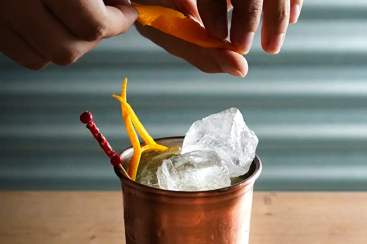 A copper cup with large ice cubes, garnished with orange peels and a red skewer, as hands squeeze an orange peel over the drink.