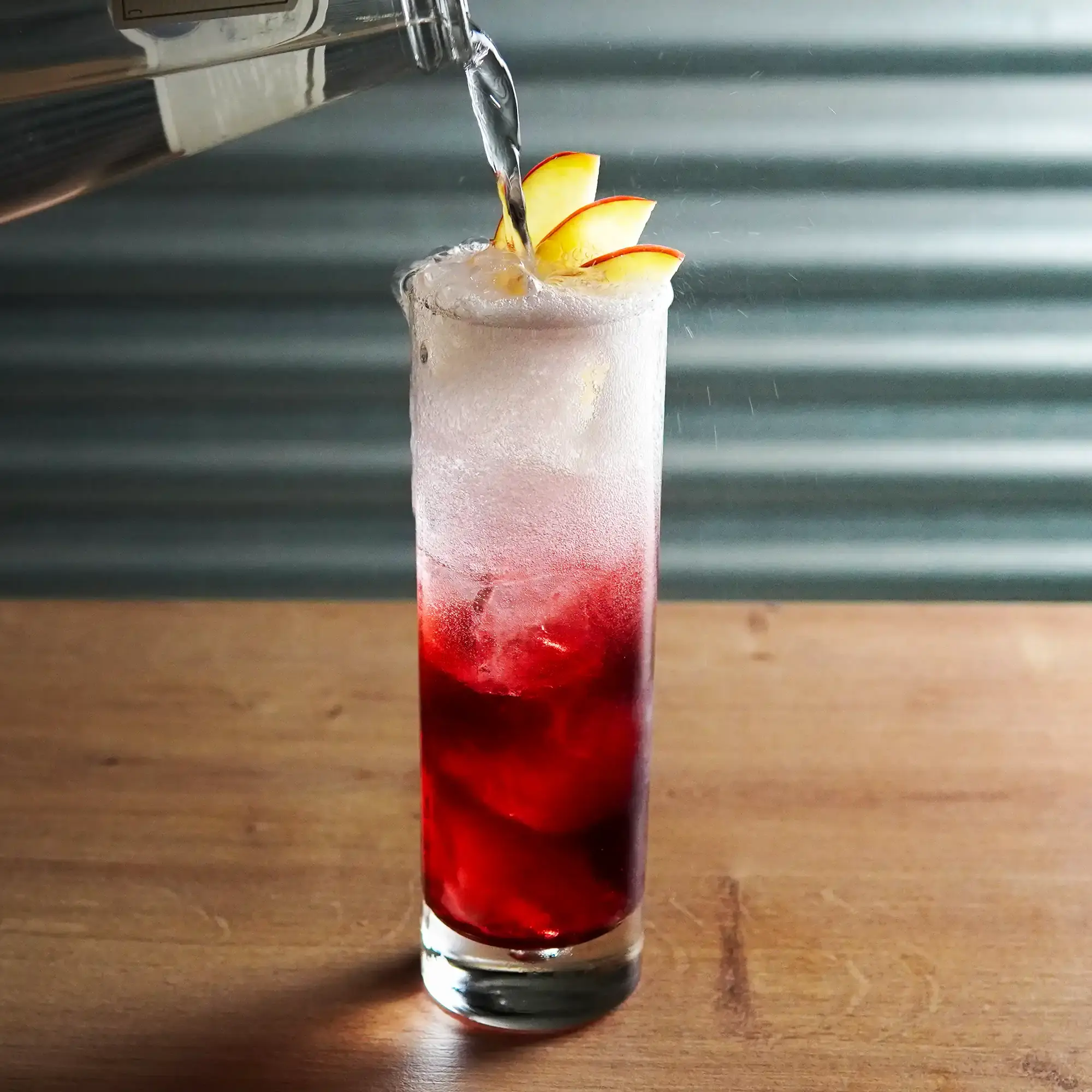 The image shows a tall, narrow cocktail glass on a light wooden surface. The drink in the glass has a vibrant color gradient: the lower part is deep red, gradually lightening toward the top, with a light foam layer on the surface. Several thin apple slices are decoratively placed along the rim, adding a fresh, fruity touch. At the top of the image, a hand is pouring a clear liquid – likely water or a clear drink – from a bottle into the glass. The liquid creates small splashes and movement on the surface, adding a dynamic element to the image. The background is plain and gray, making the drink’s colors and apple slices stand out prominently.