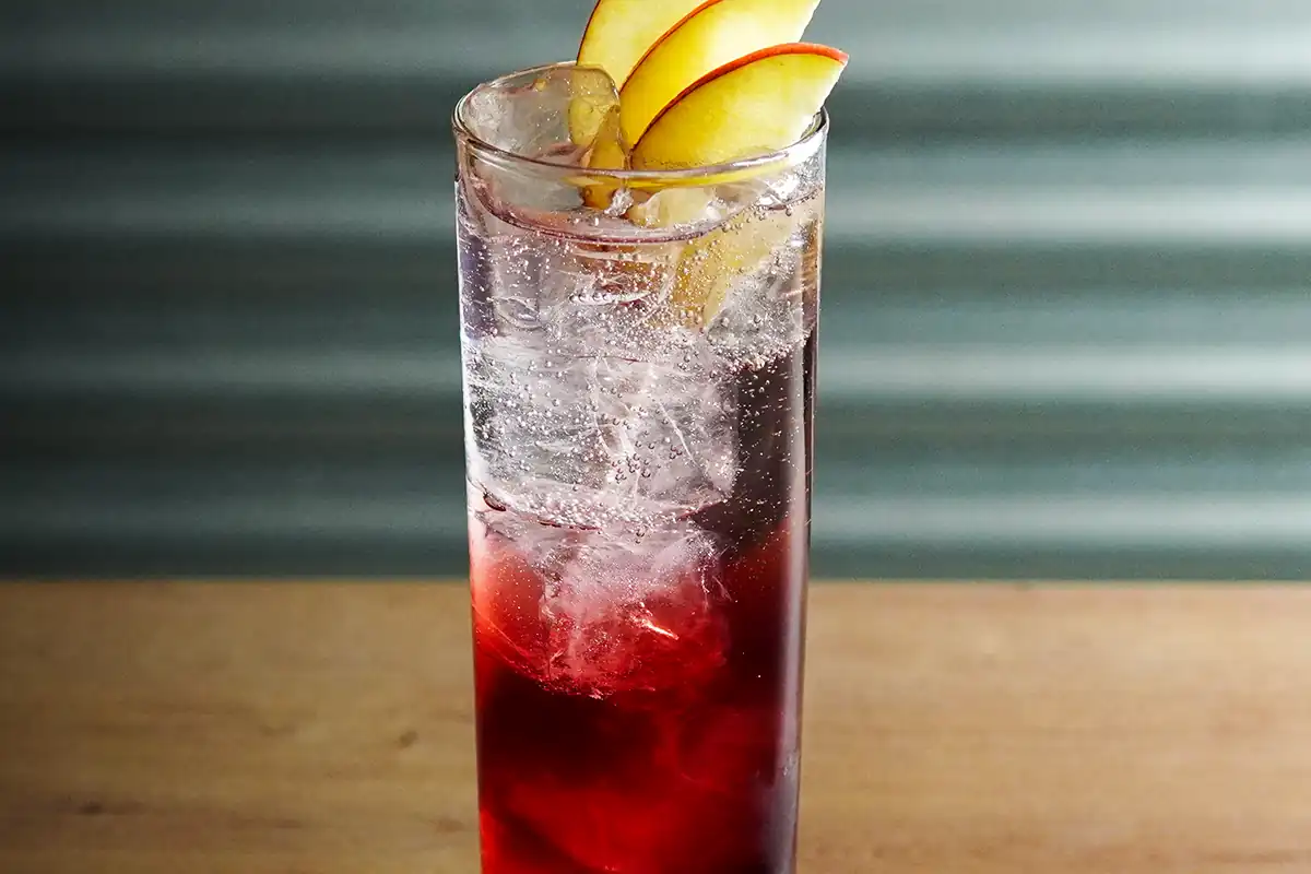 A tall glass filled with a red and clear layered drink, garnished with ice cubes and thin apple slices on top.