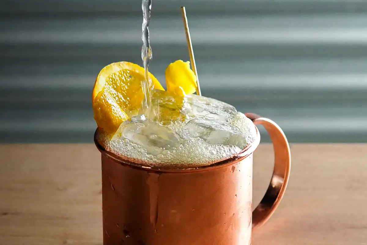 A copper mug filled with a foamy, bubbly drink, garnished with orange slices and a lemon twist, as liquid is being poured in.