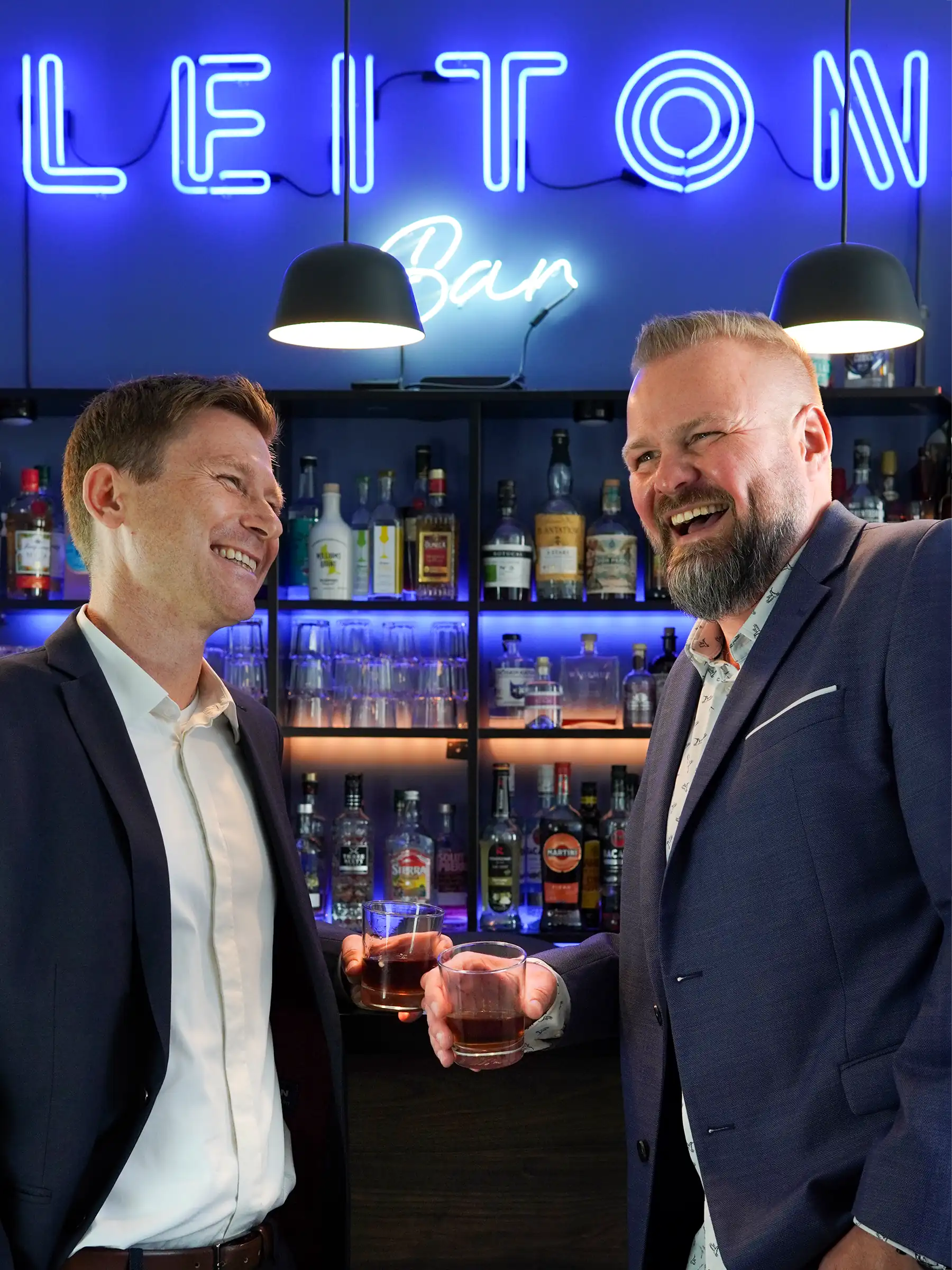 Two men in business attire holding drinks and laughing in front of a bar setup with a neon "LEITON Bar" sign, illuminated bottles, and glasses in the background.