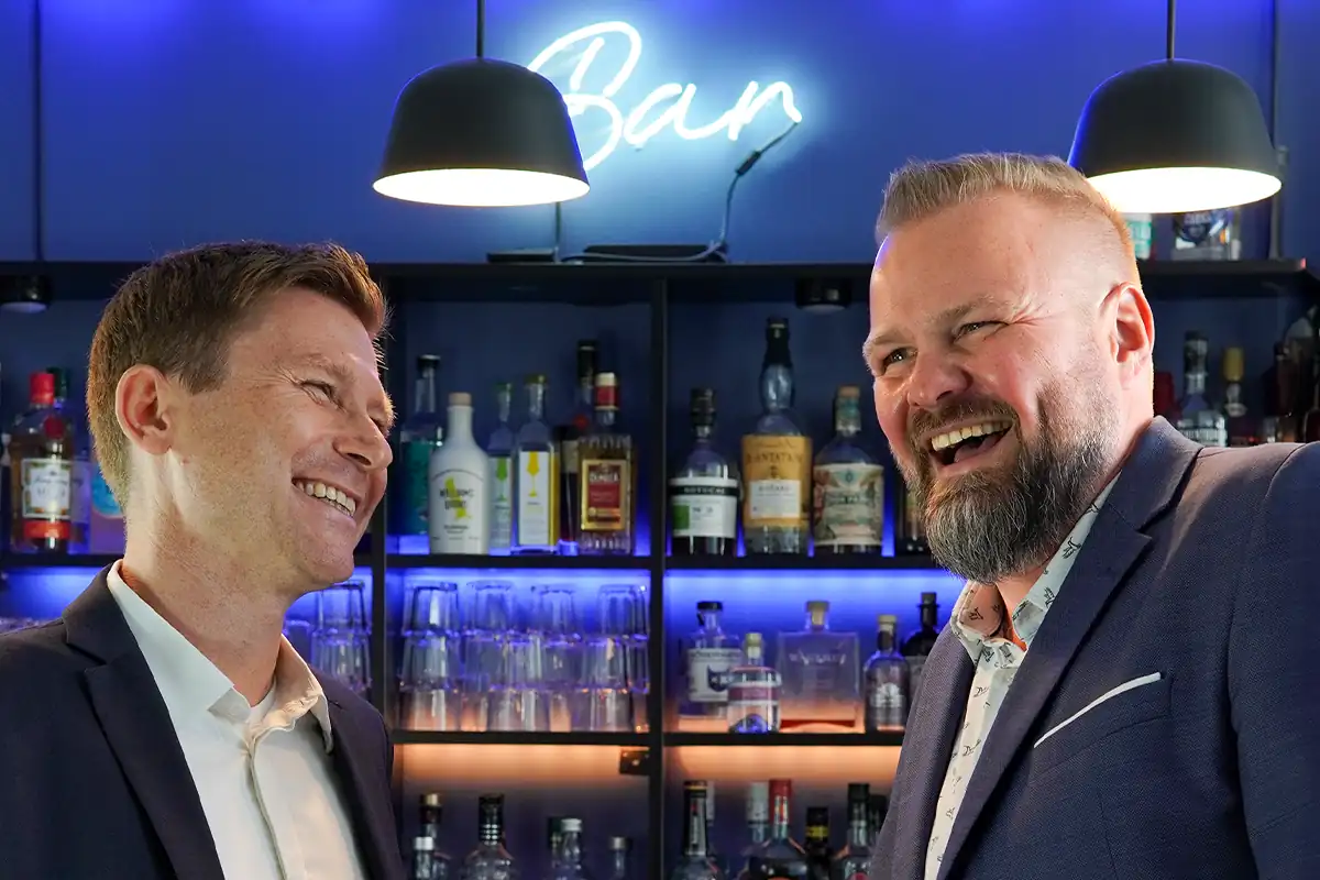 Two men in business attire laughing together in front of a bar setup with illuminated bottles and a neon "Bar" sign in the background.