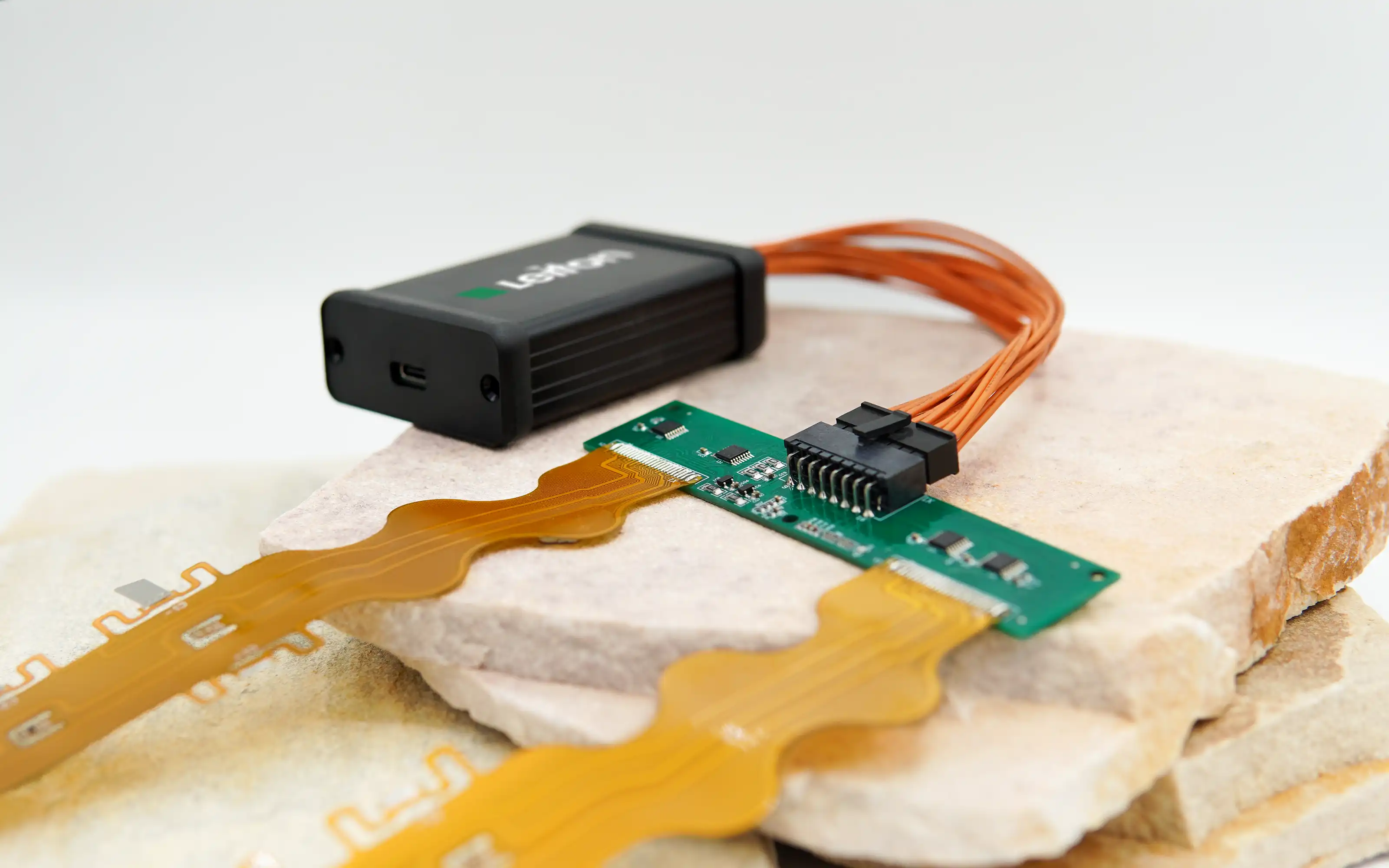 A flexible printed circuit board (PCB) with orange connectors attached to a green adapter board, connected to a black Leiton device, placed on stacked stone slabs.