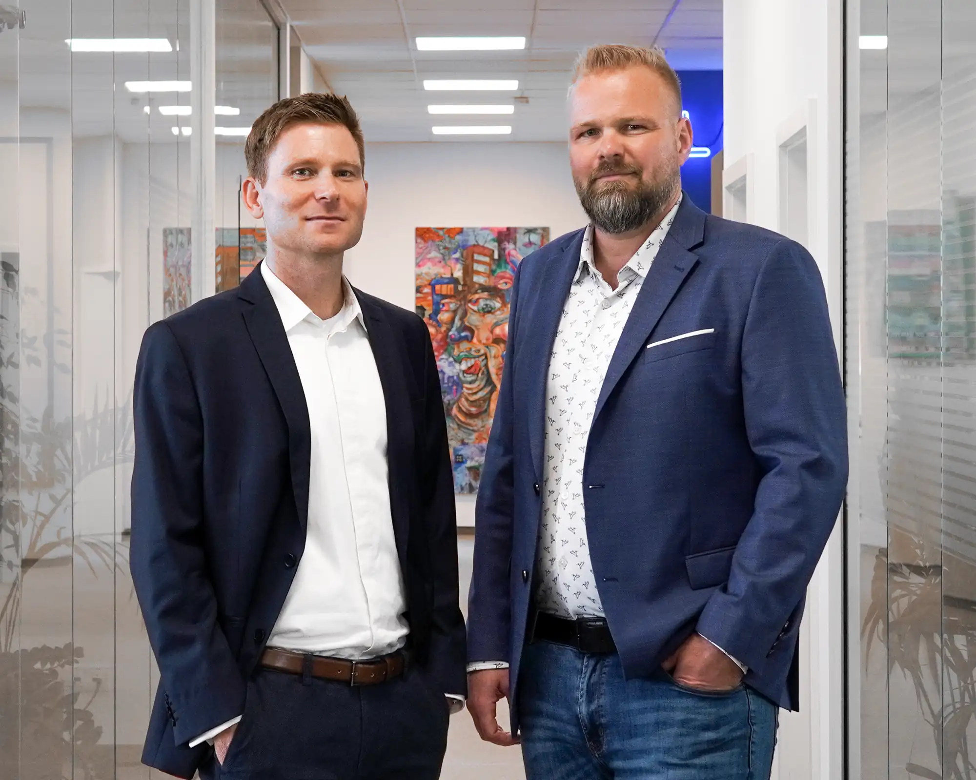 Two men in business attire stand confidently in a modern office hallway with contemporary artwork visible on the wall behind them.